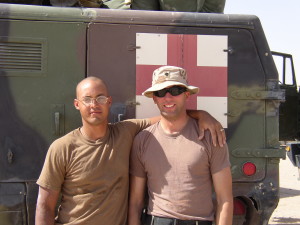 Two soldiers stand in front of a n ambulance bearing a red cross.